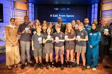 A group of children standing with Steve Harvey, in front of a screen that says "A Call from Space"