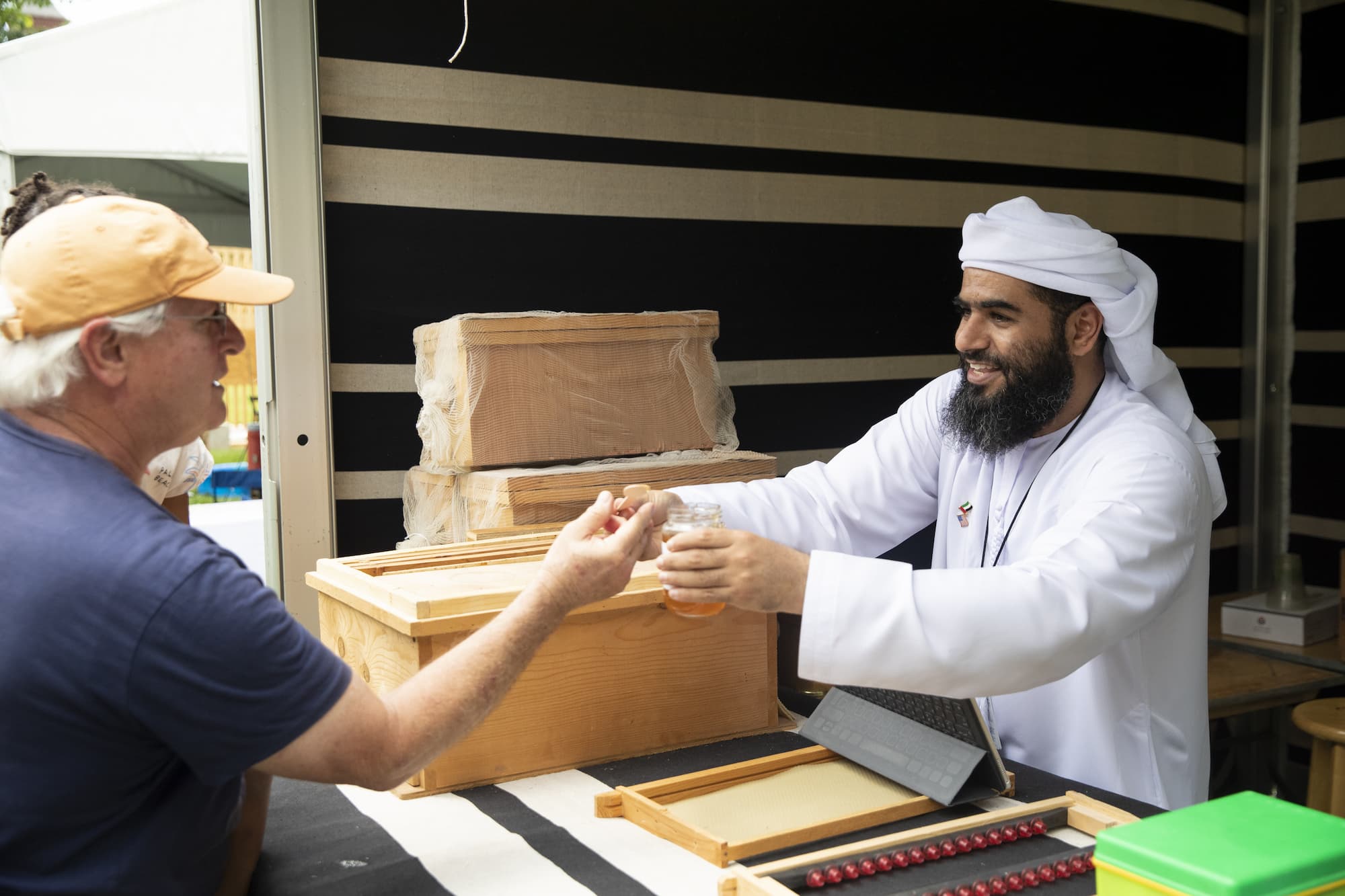 Manea Al Kaabi passing a honey tasting sample to a visitor wearing a baseball cap