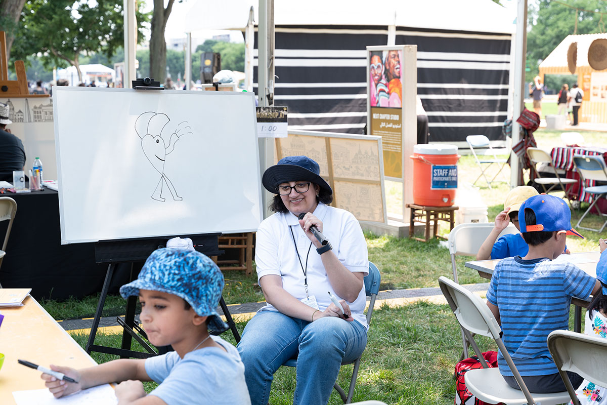 Woman wearing a blue bucket hat, instructing a group of children on how to draw a cartoon