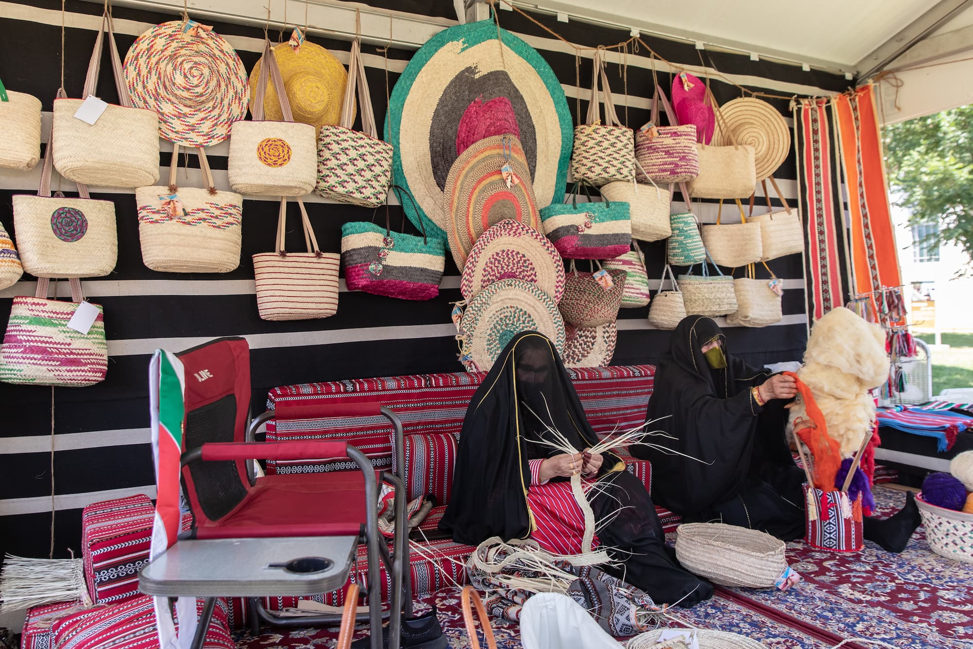 Two women sitting on a carpeted floor, weaving baskets, in front of a large display of completed baskets