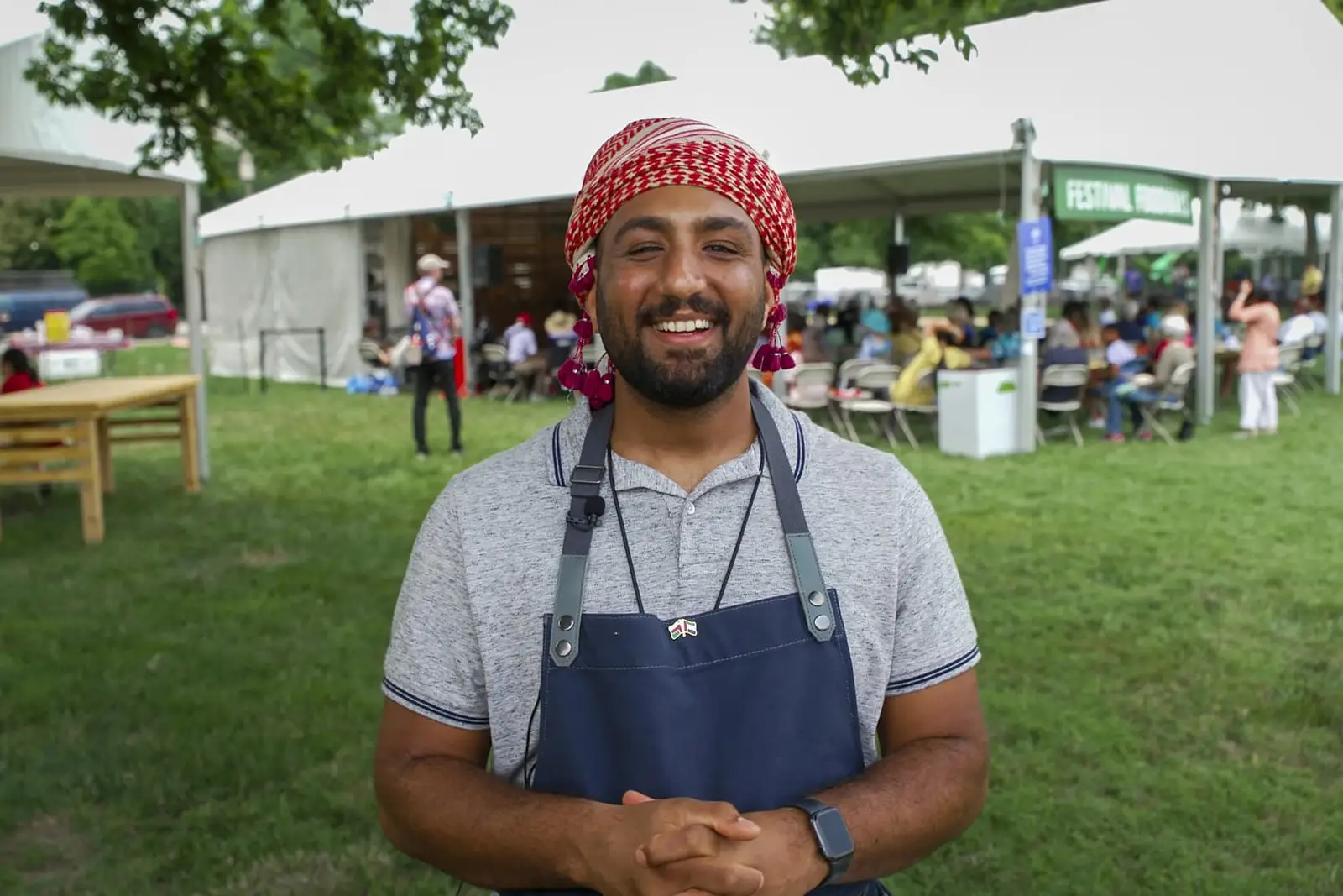 Smiling man wearing a red head covering and a blue chef's apron standing in front of a tent with a crowd gathered underneath