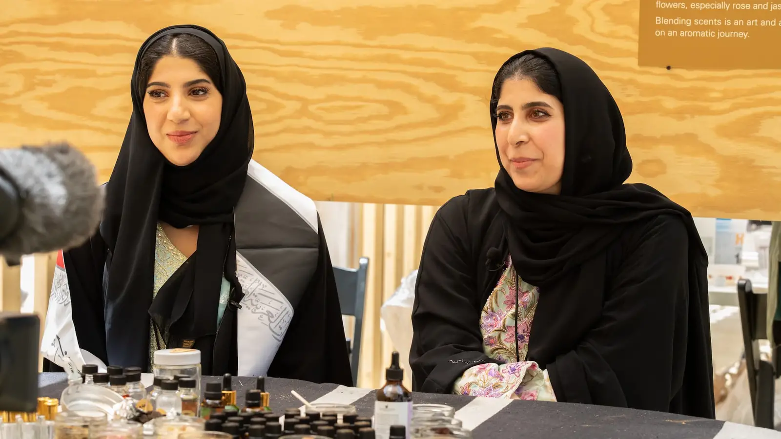 Two women in black headscarves sitting next to each other, with perfume bottles and essential oils on the table in front of them