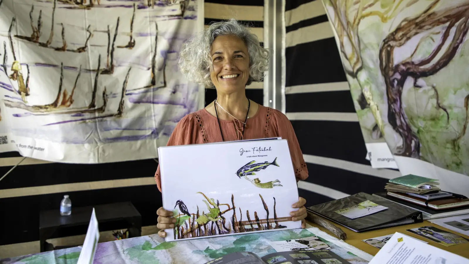 Woman with grey hair holding up a painting, standing in front of hand-painted tapestries