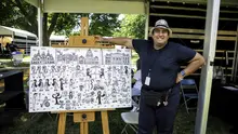 A smiling man standing next to a big canvas filled with black ink cartoon drawings