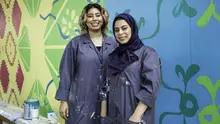 Two women in navy painters smocks standing in front of a colorful abstract mural