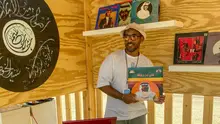 Man wearing a hat and sunglasses holding a record album, standing in front of two shelves displaying records