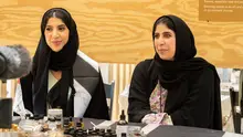 Two women in black headscarves sitting next to each other, with perfume bottles and essential oils on the table in front of them
