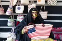 Woman in black headscarf holding an American flag printed woven bag, in front of a display wall with other handmade woven bags and baskets